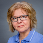 Ältere Frau mit blonden Haaren, schwarzer Brille und blauem Hemd blickt neutral in die Kamera vor grauem Hintergrund.