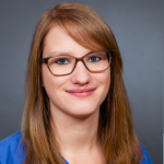 Frau mit glattem hellbraunem Haar und Brille, lächelt in einem blauen Oberteil vor grauem Hintergrund in die Kamera.