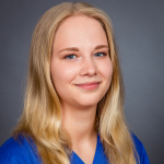 Blonde Frau im blauen Oberteil lächelt sanft vor einem schlichten grauen Hintergrund in einem professionellen Porträt.