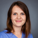 Lächelnde Frau mit glattem braunem Haar und blauen Augen, in einem hellblauen Oberteil, posiert vor einem grauen Hintergrund.