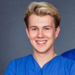 Junge Person mit kurzen blonden Haaren in blauer OP-Kleidung lächelt in die Kamera, schlichter grauer Hintergrund.