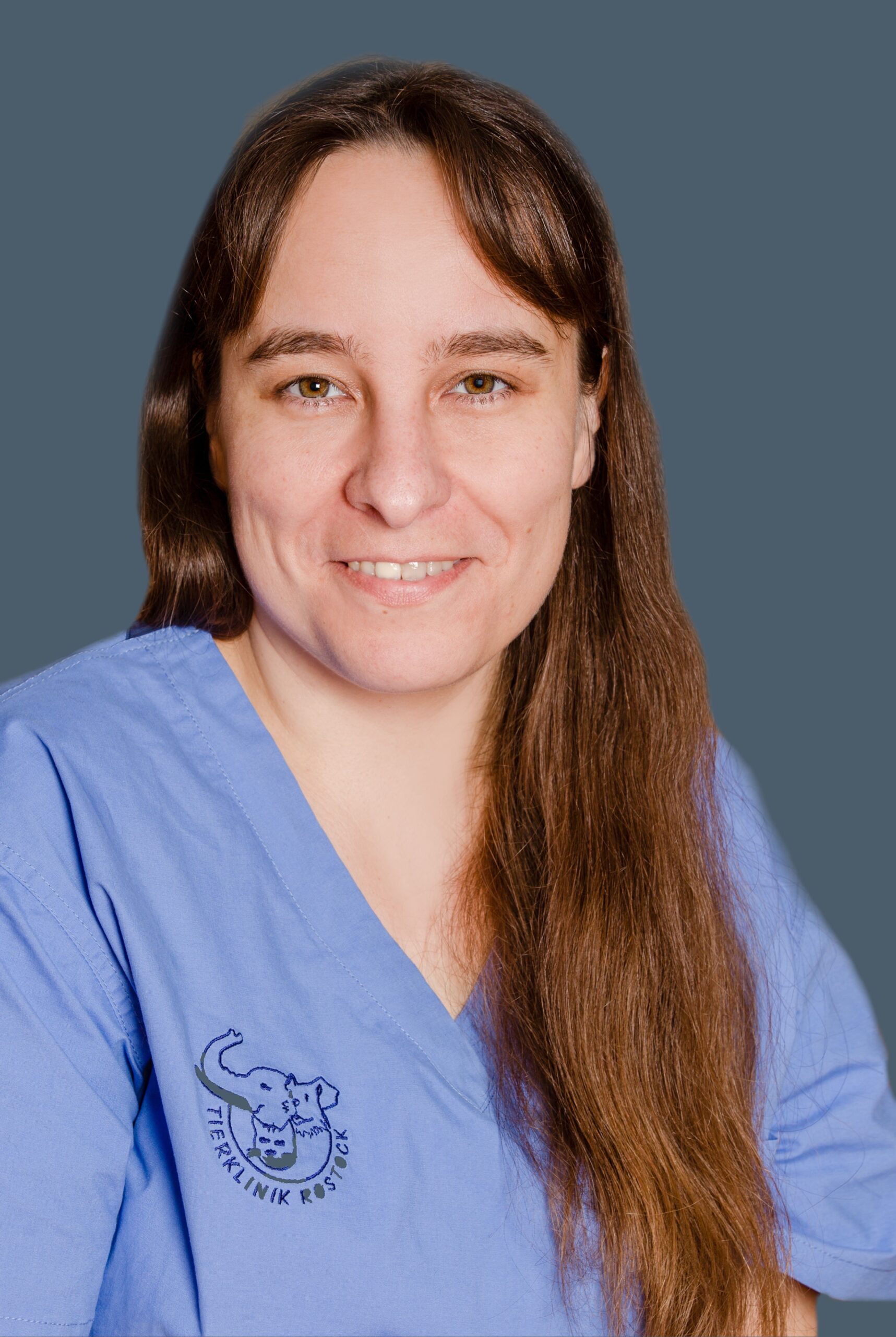 Mariana Kohn, lächelnd mit langen braunen Haaren, trägt einen blauen Kittel aus der Tierklinik vor einem schlichten grauen Hintergrund.