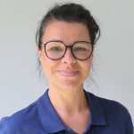 Frau mit dunklem, zurückgebundenem Haar, großer runder Brille und marineblauem Kragenhemd, lächelnd vor einem schlichten hellen Hintergrund.
