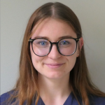 Junge Frau mit langen braunen Haaren, runder Brille und marineblauem Oberteil lächelt sanft vor einem schlichten hellen Hintergrund.