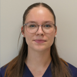 Junge Frau mit Brille, Perlenohrringen und marineblauem Oberteil lächelt leicht vor einem schlichten hellen Hintergrund.
